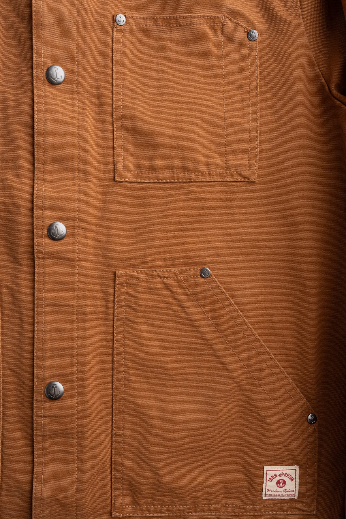 Brown canvas jacket with metal buttons, two front pockets, and a small label at the bottom right corner.