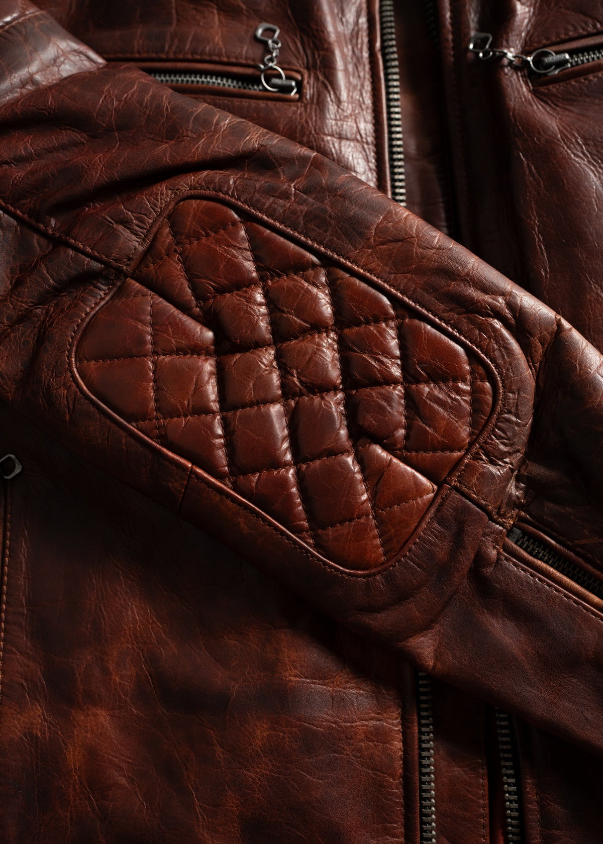 Close-up of a brown leather jacket with quilted elbow patch and zippers, highlighting texture and craftsmanship.