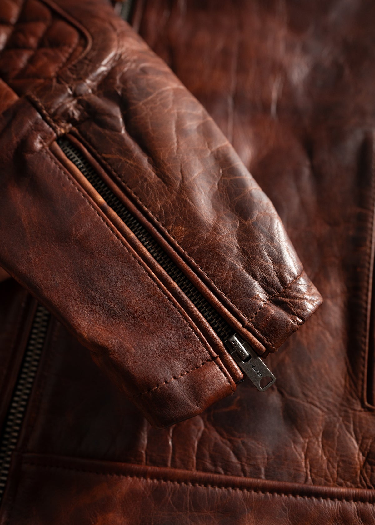 Close-up of a brown leather jacket sleeve with visible stitching and a metal zipper, highlighting texture and craftsmanship.