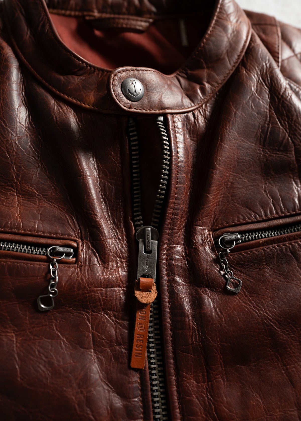 Close-up of a brown leather jacket with a zipper and button detail, highlighting texture and craftsmanship in soft lighting.