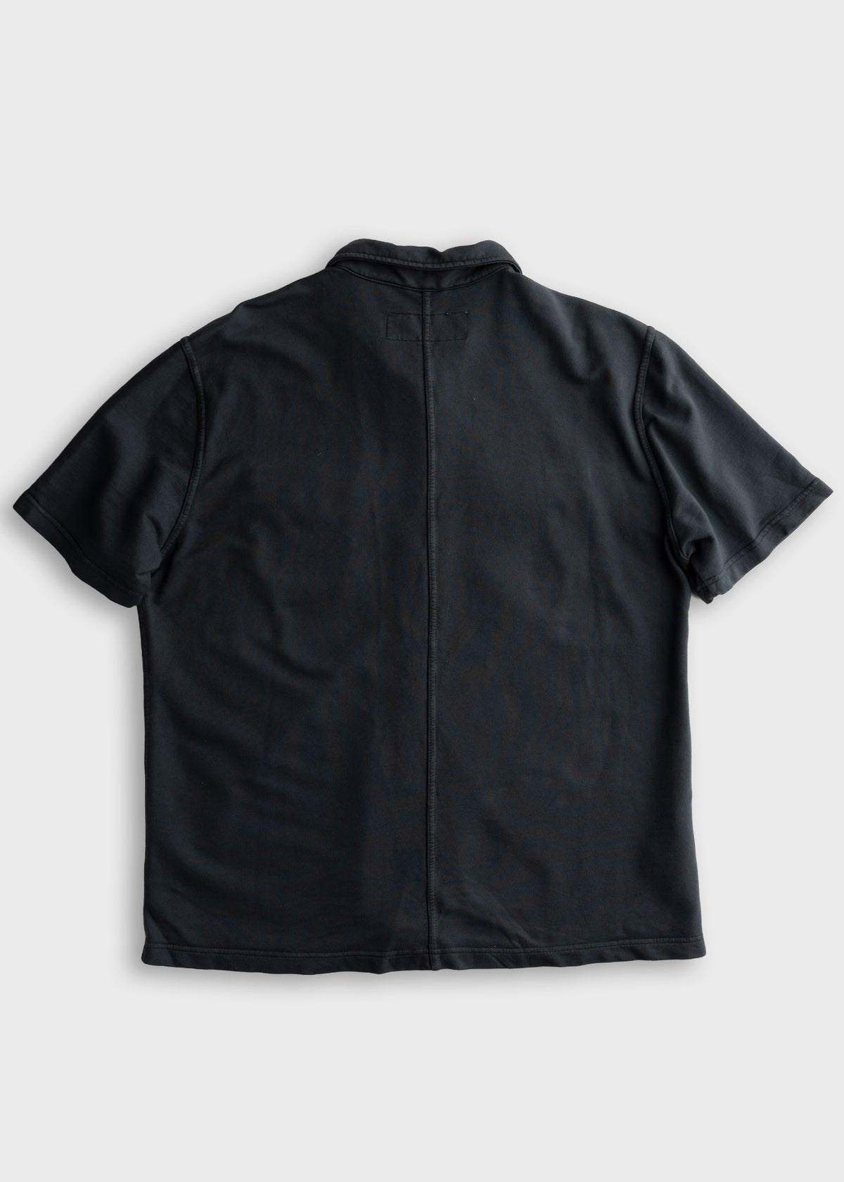 Back view of a black short-sleeve shirt on a plain background, highlighting its simple design and straight seam down the center.