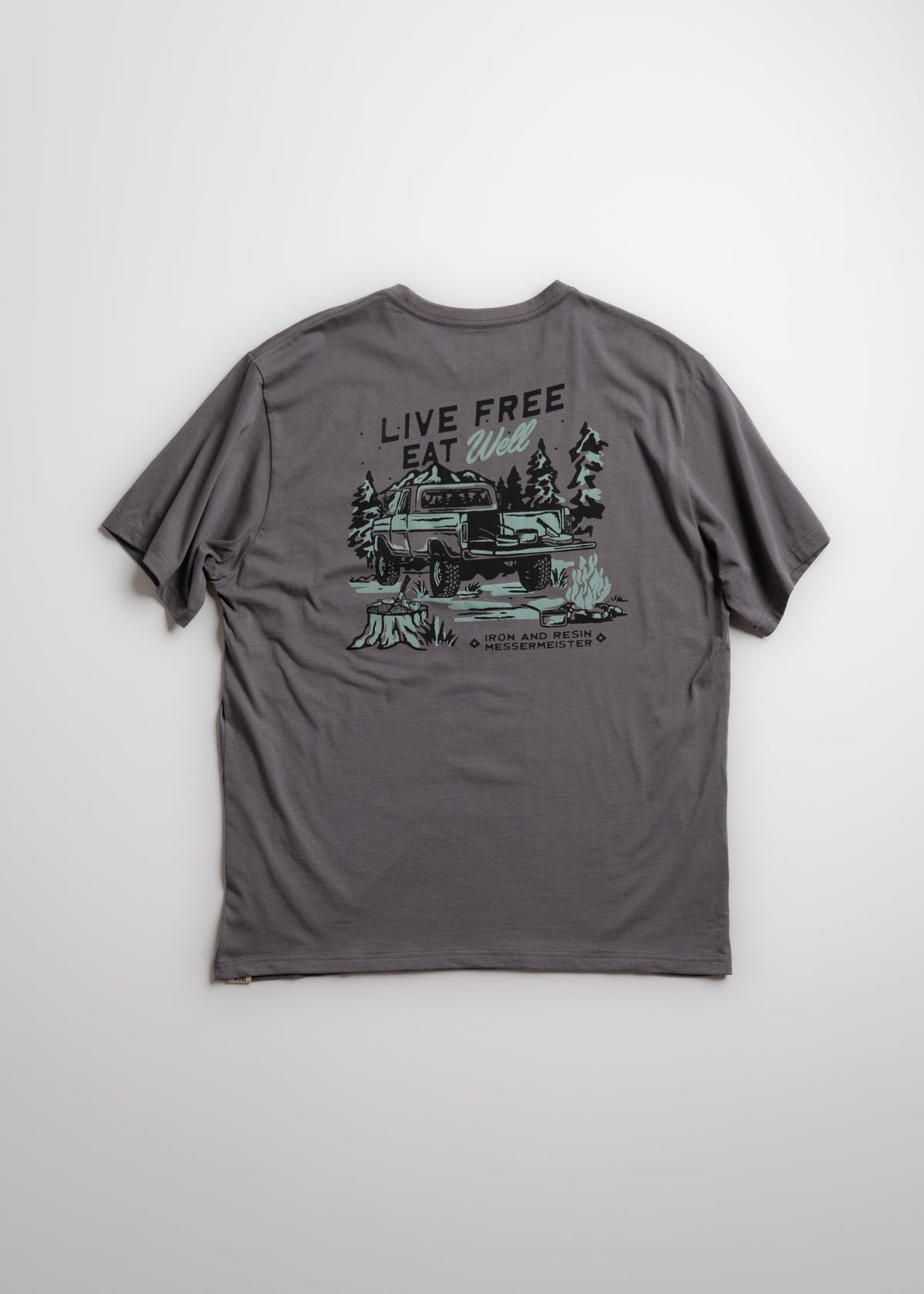 Gray T-shirt with "Live Free, Eat Well" slogan and truck graphic in forest setting. Casual outdoor adventure theme.