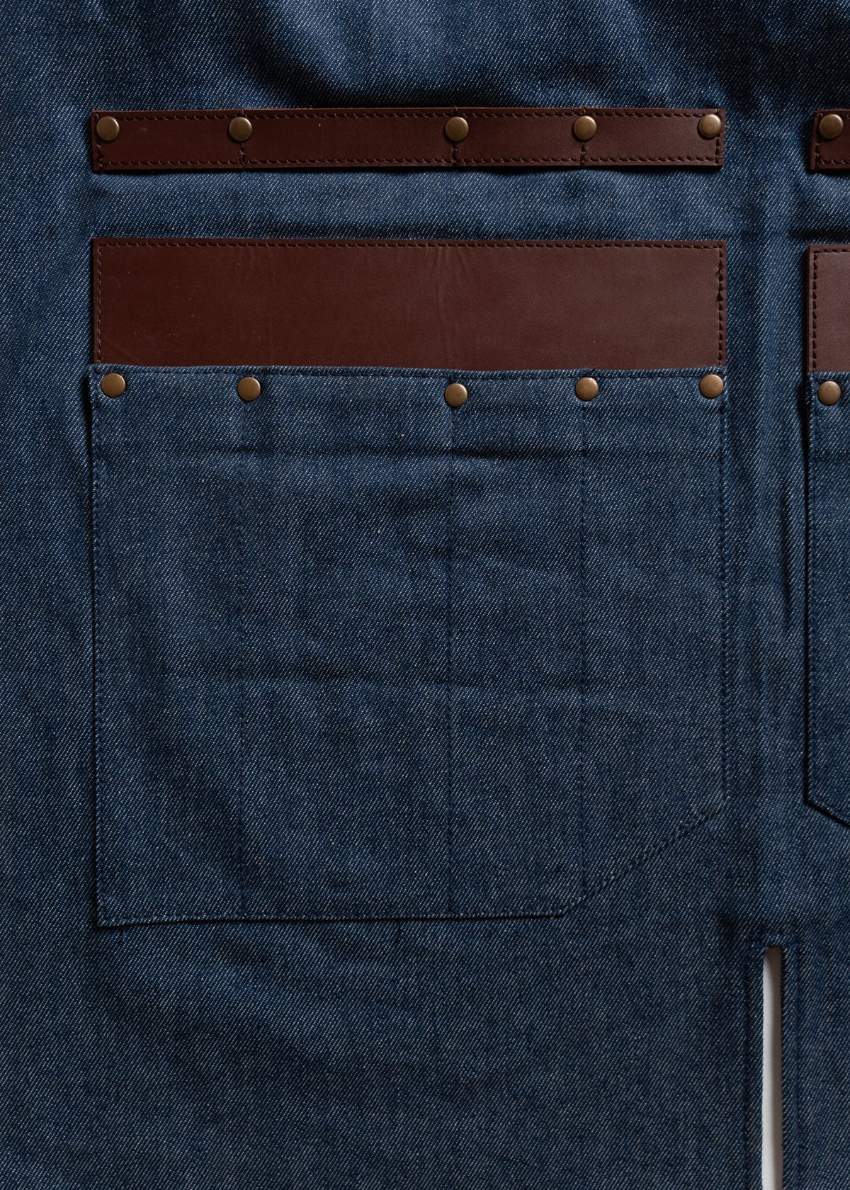 Denim apron with brown leather accents and rivets, featuring large front pockets. Durable and stylish design for work or craft.