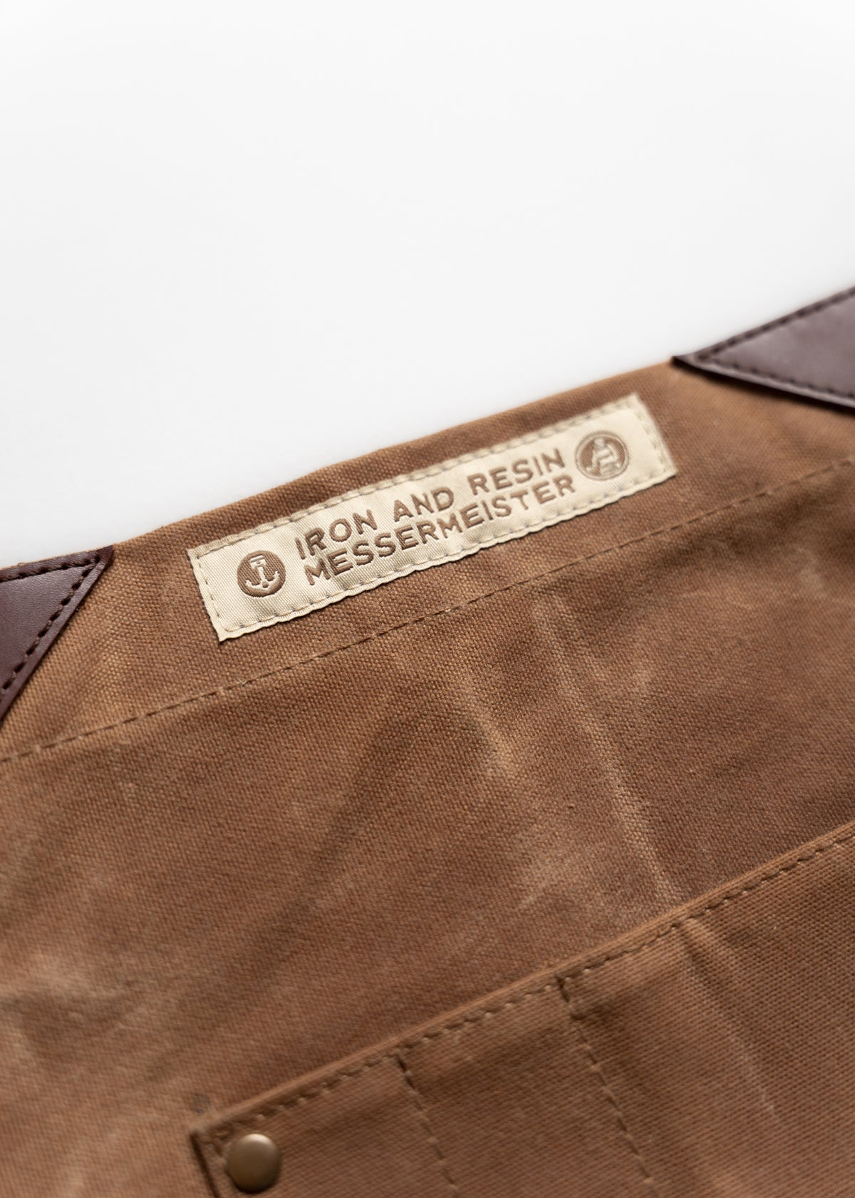 Brown fabric apron with "Iron and Resin Messermeister" label, featuring leather corners and a riveted pocket for durability.