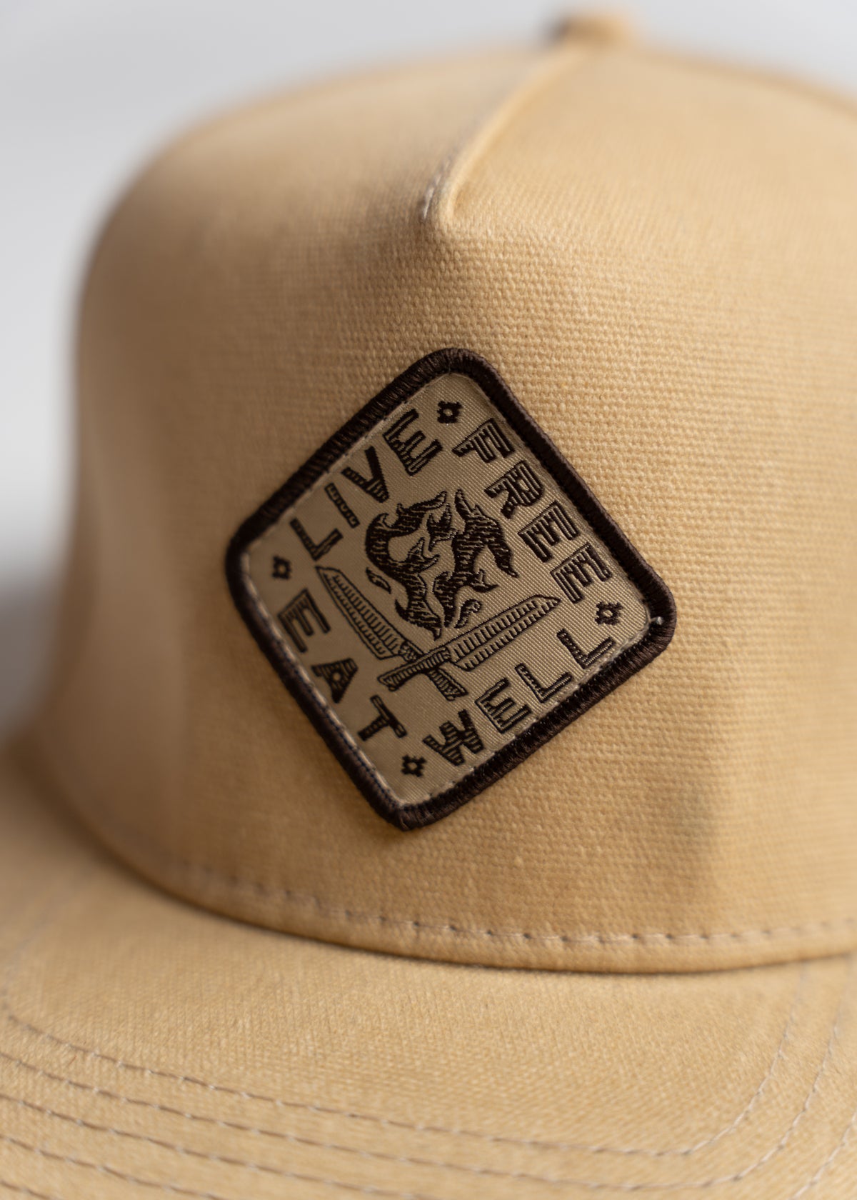 Beige cap with a diamond-shaped patch reading "Live Free, Eat Well" in bold black text, featuring a small graphic design.