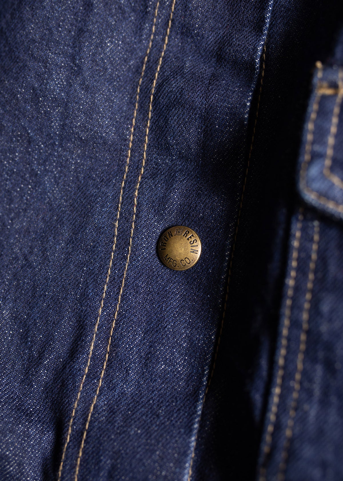Close-up of denim fabric with visible stitching and a brass button engraved with "MFG. CO." on the right side.