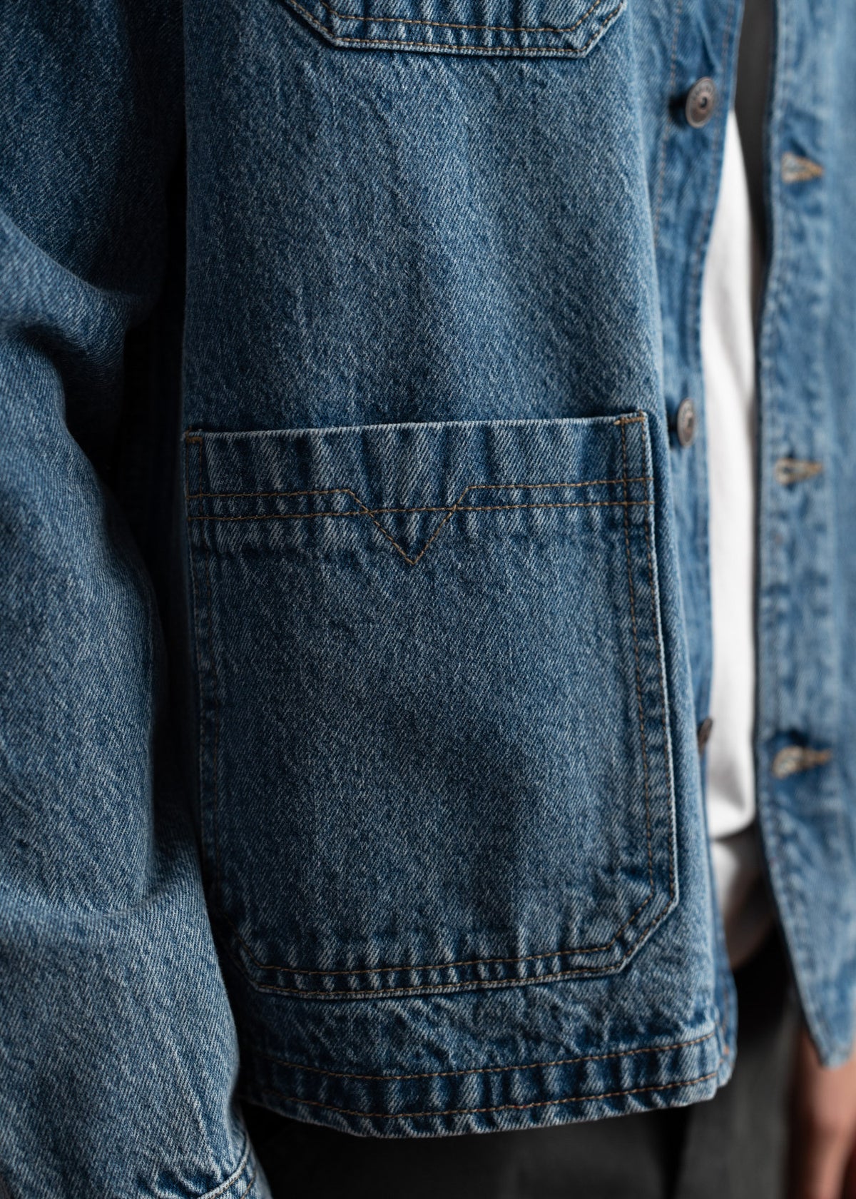 Close-up of a blue denim jacket with visible stitching and a pocket, worn over a white shirt.