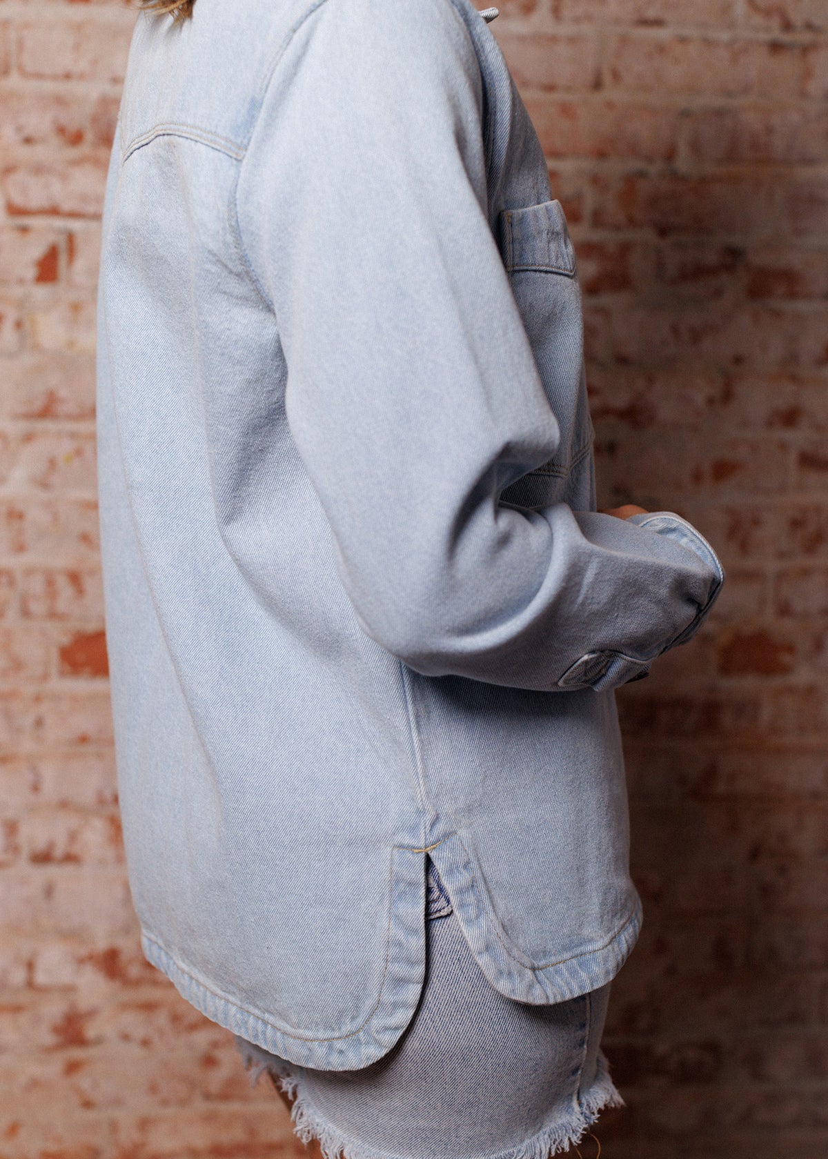 A person in a light denim jacket and shorts stands against a rustic brick wall, showcasing casual style and texture contrast.
