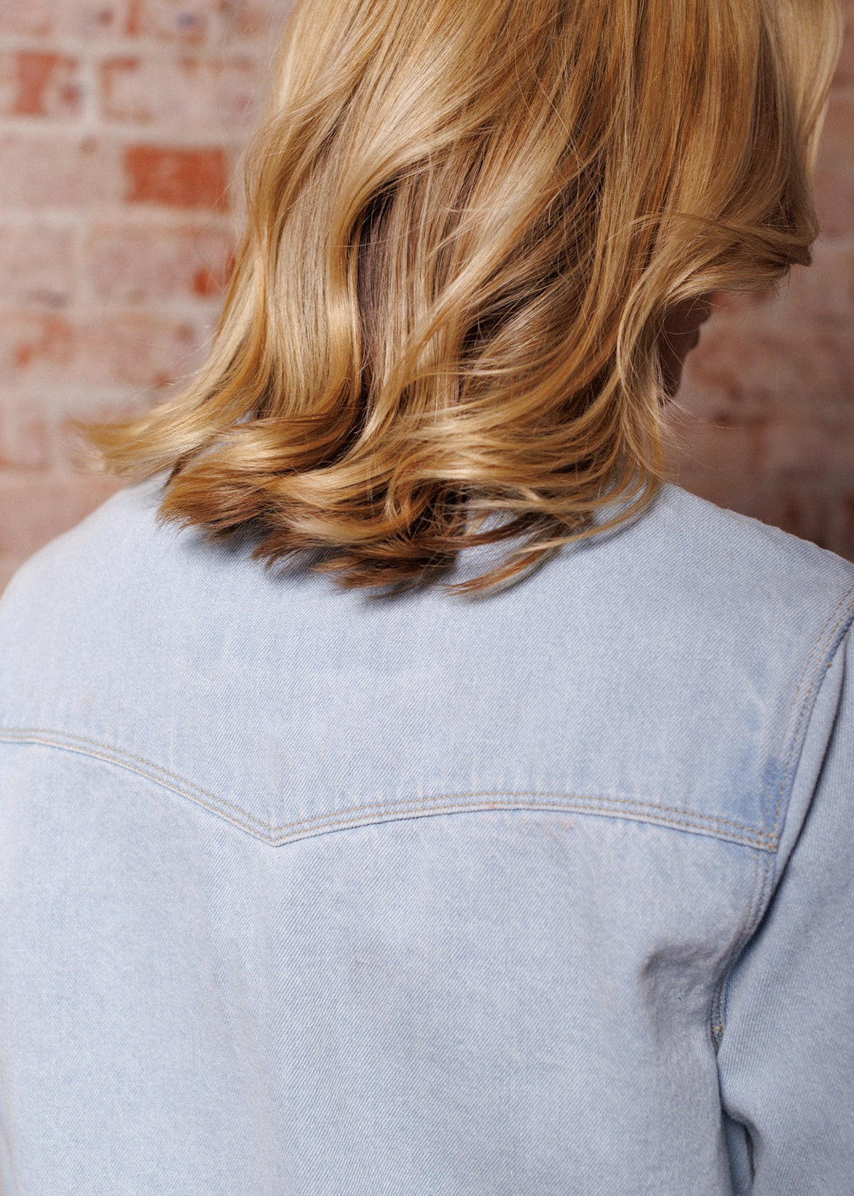 Blonde hair cascades over a light denim jacket, set against a rustic brick wall, capturing a casual and stylish moment.