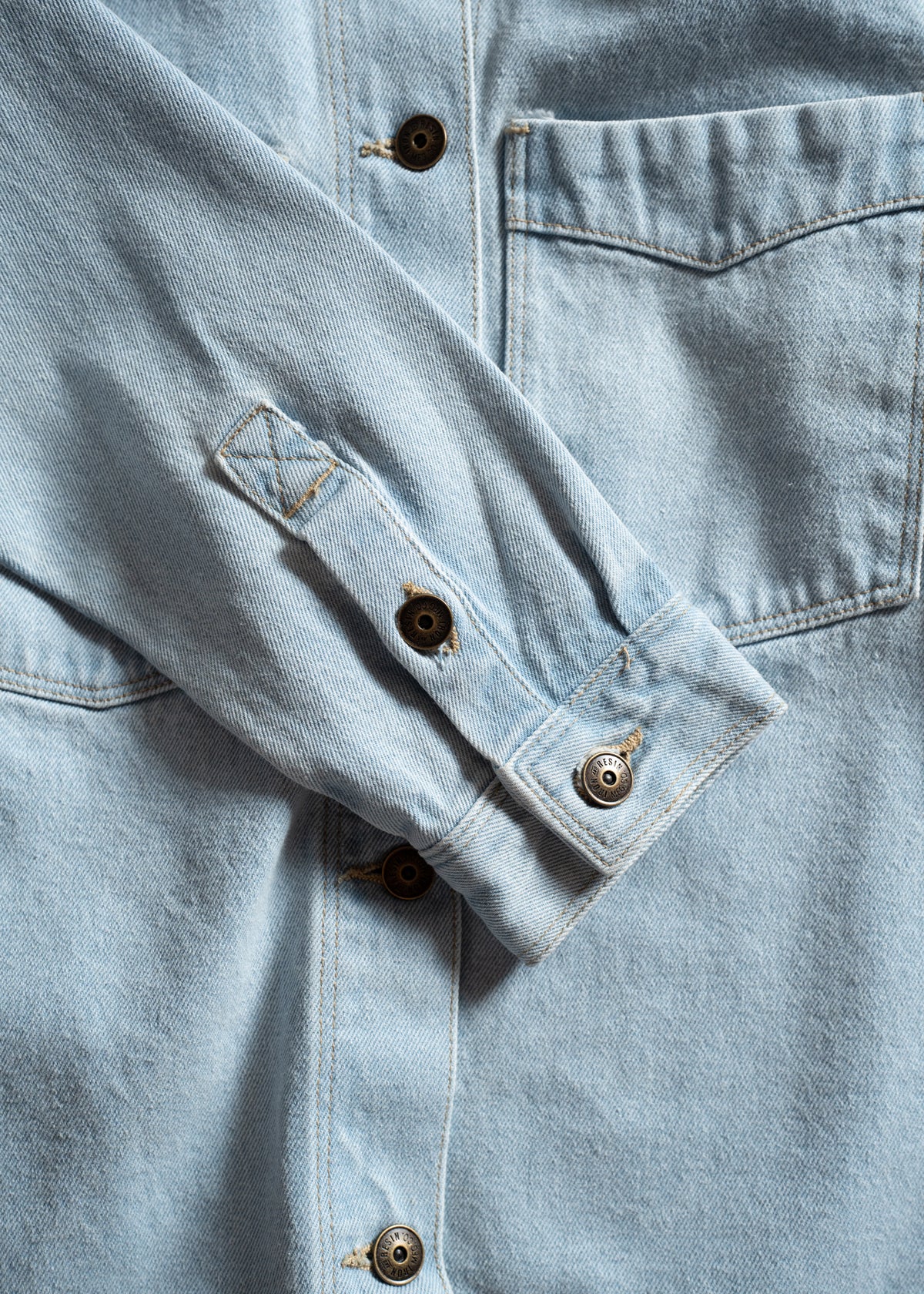 Close-up of a light blue denim shirt with metal buttons and a chest pocket, showcasing its texture and stitching details.
