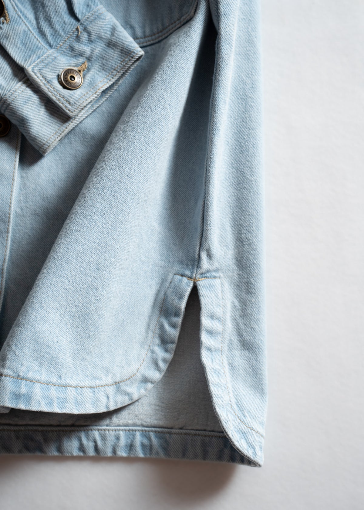 Close-up of a light blue denim shirt's hem and buttoned cuff, showcasing detailed stitching and fabric texture.