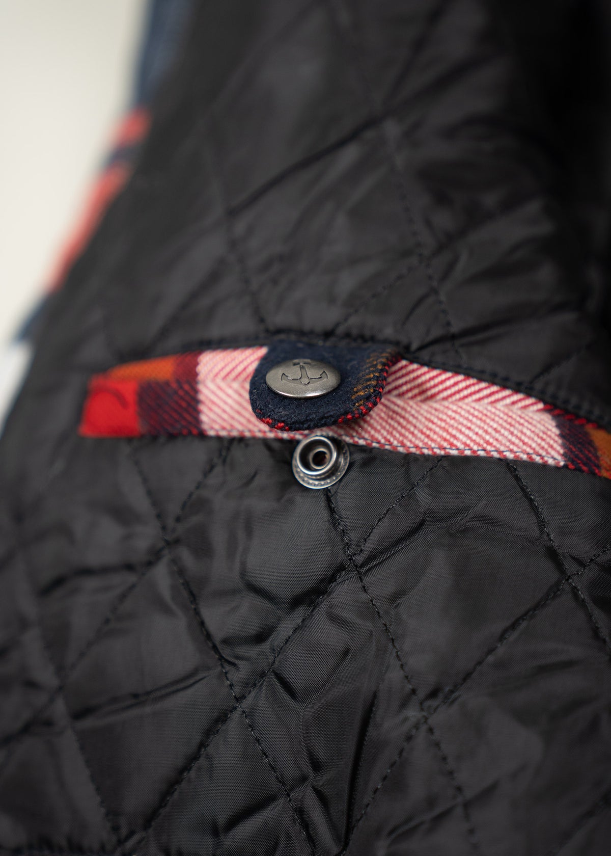 Close-up of a quilted black fabric with a red plaid trim and a metal button featuring an anchor design.