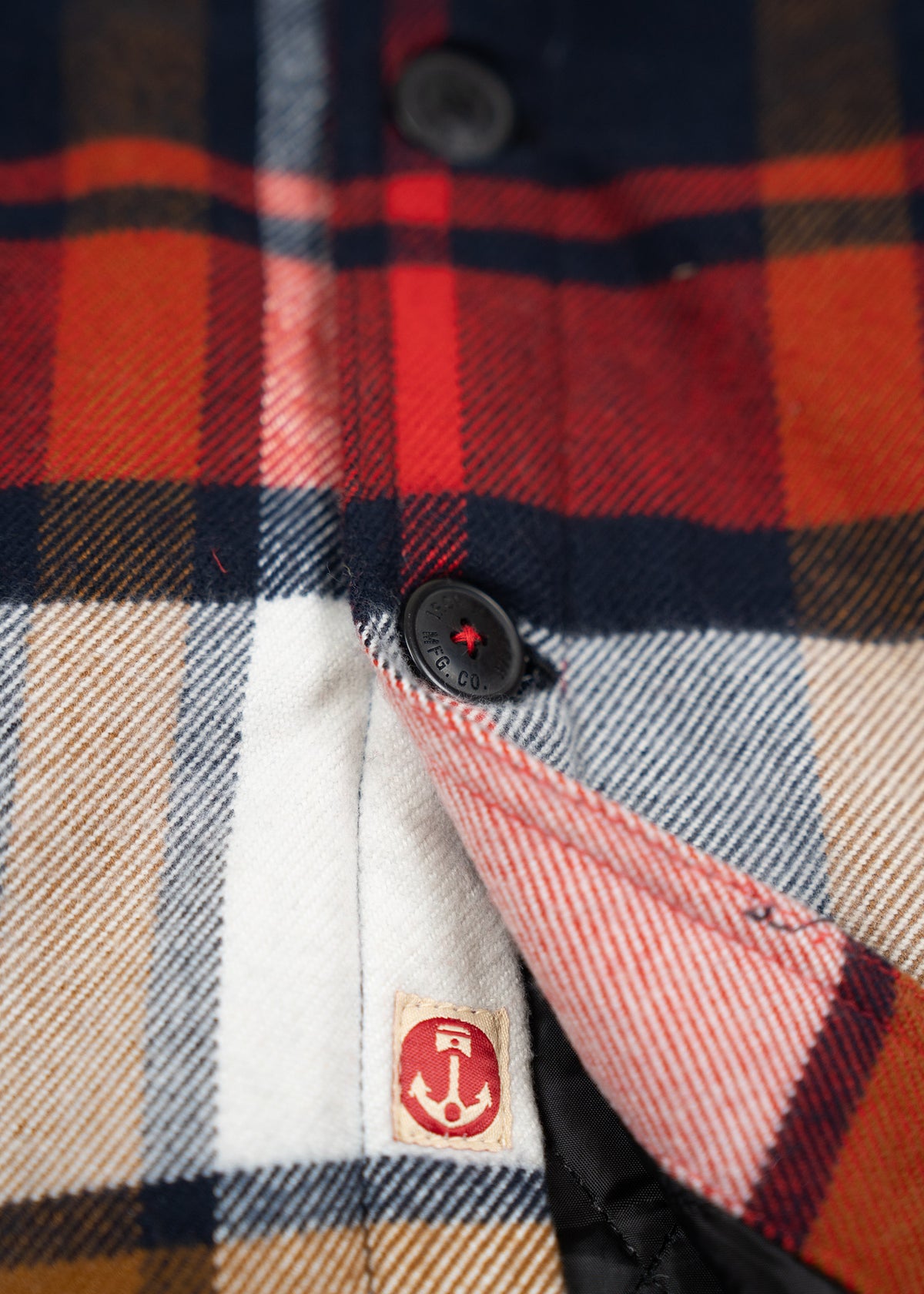 Close-up of a red, navy, and white plaid shirt with black buttons and an anchor logo tag on the inner seam.