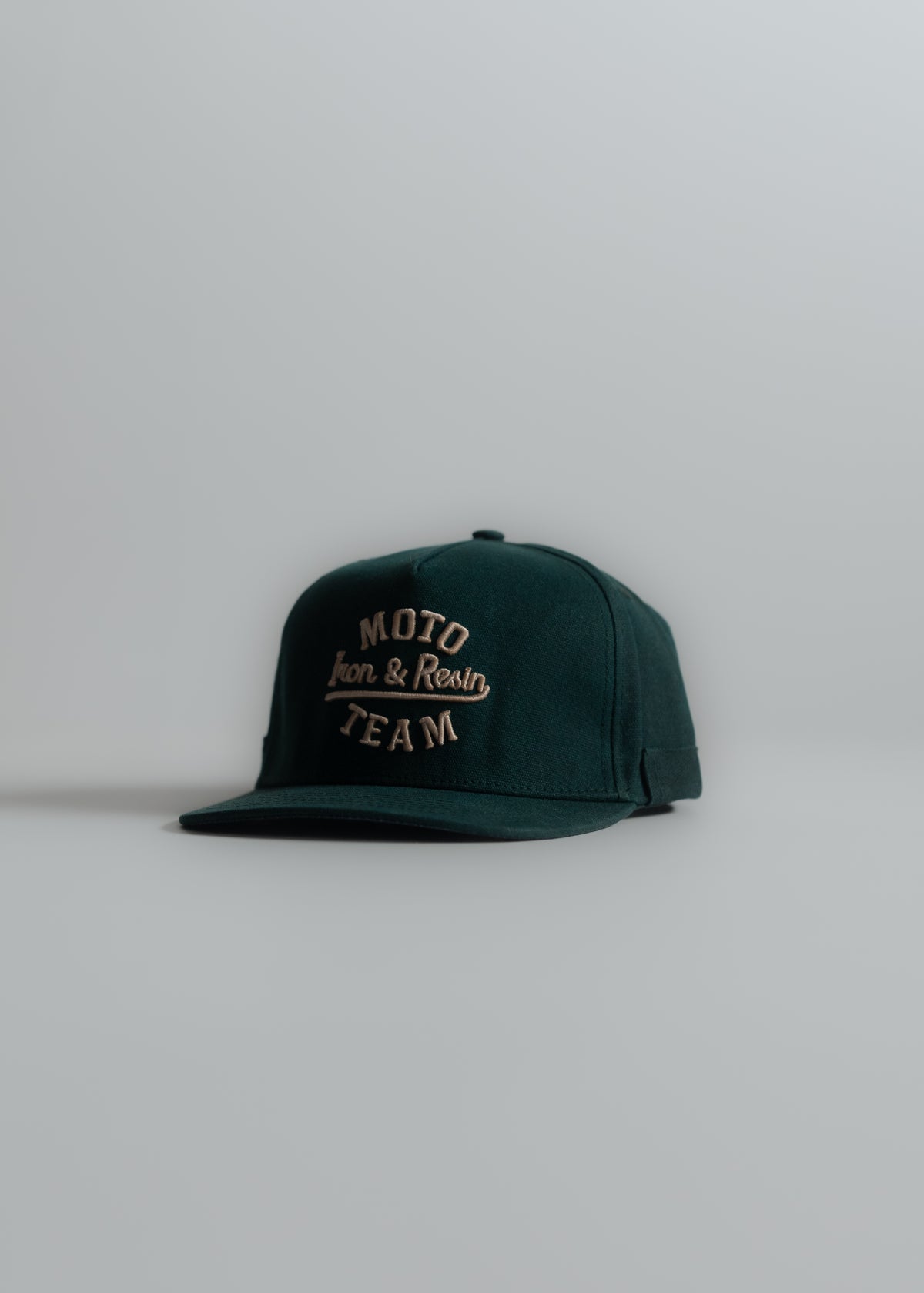 Green cap with "Moto Iron & Resin Team" embroidered in beige, placed on a light gray surface, angled slightly to the left.