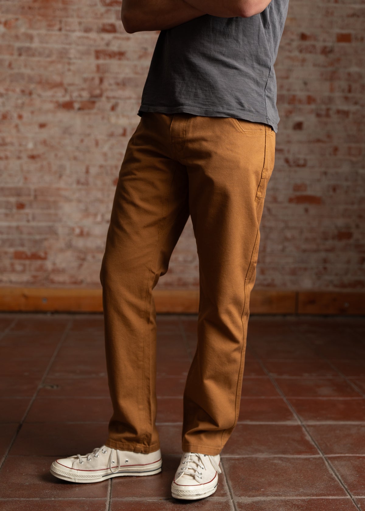 Man in a gray shirt and brown pants stands with arms crossed, wearing white sneakers on a brick floor with a brick wall backdrop.