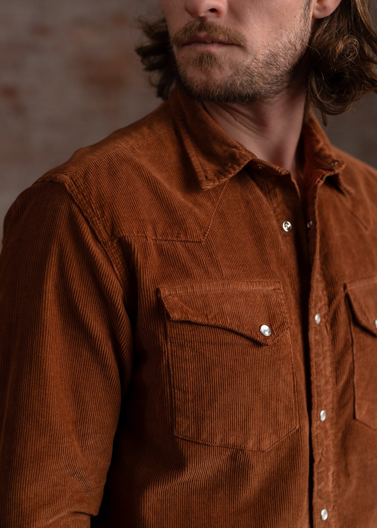 Man in a brown corduroy shirt with buttoned pockets stands against a blurred brick wall, looking to the side with a neutral expression.