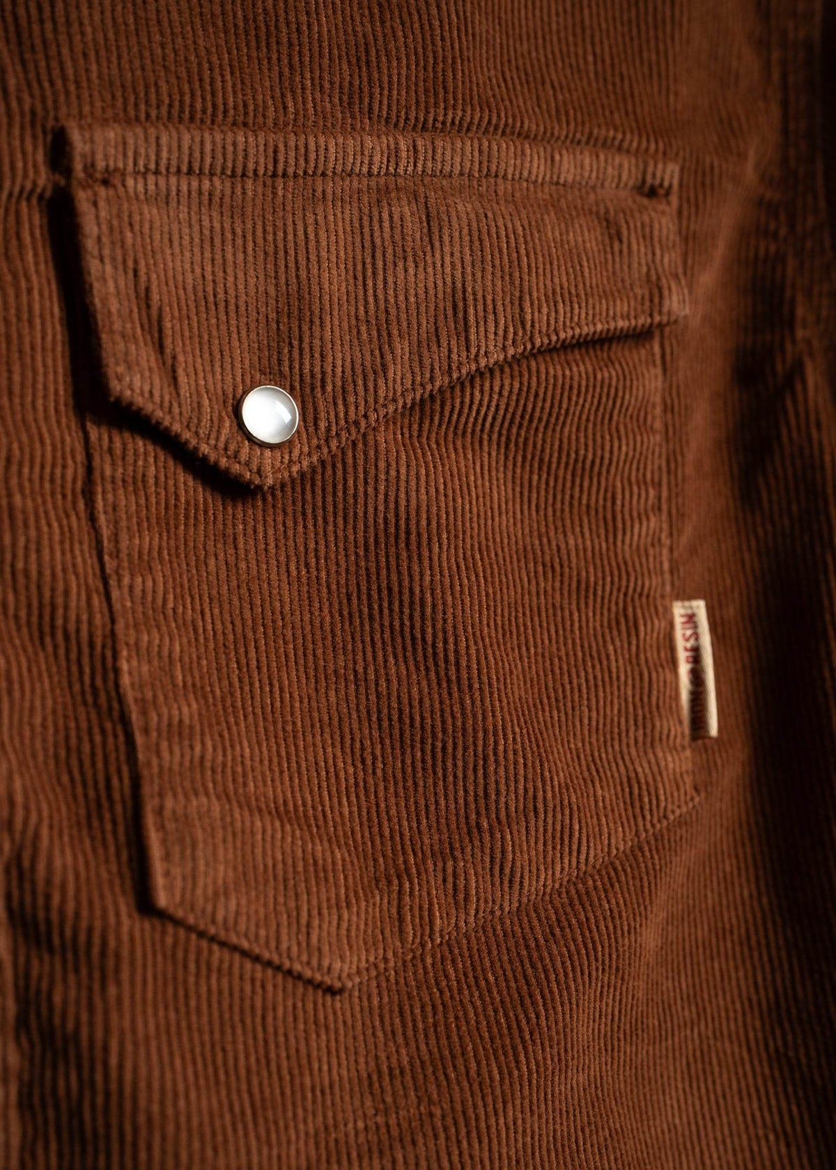 Close-up of a brown corduroy shirt pocket with a metal button, showcasing the fabric's texture and a small tag on the side.