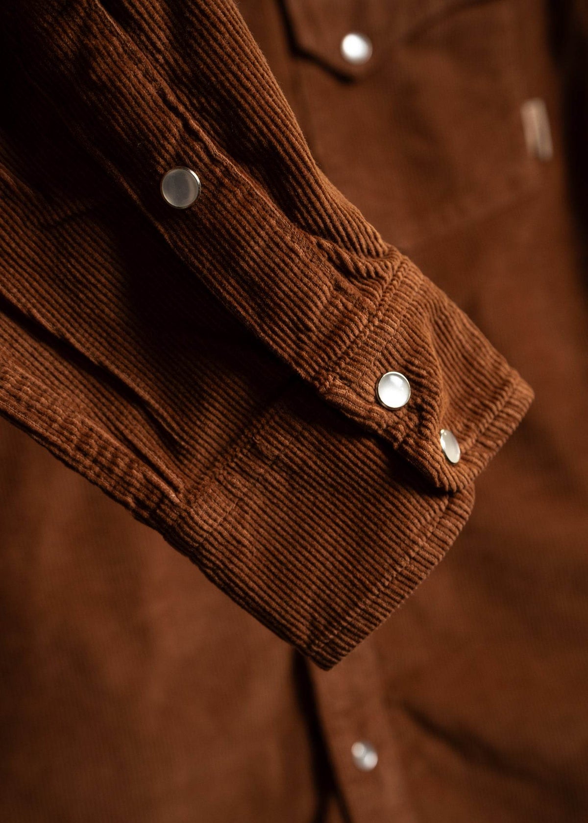 Close-up of a brown corduroy jacket sleeve with silver snap buttons, highlighting the textured fabric and detailed stitching.