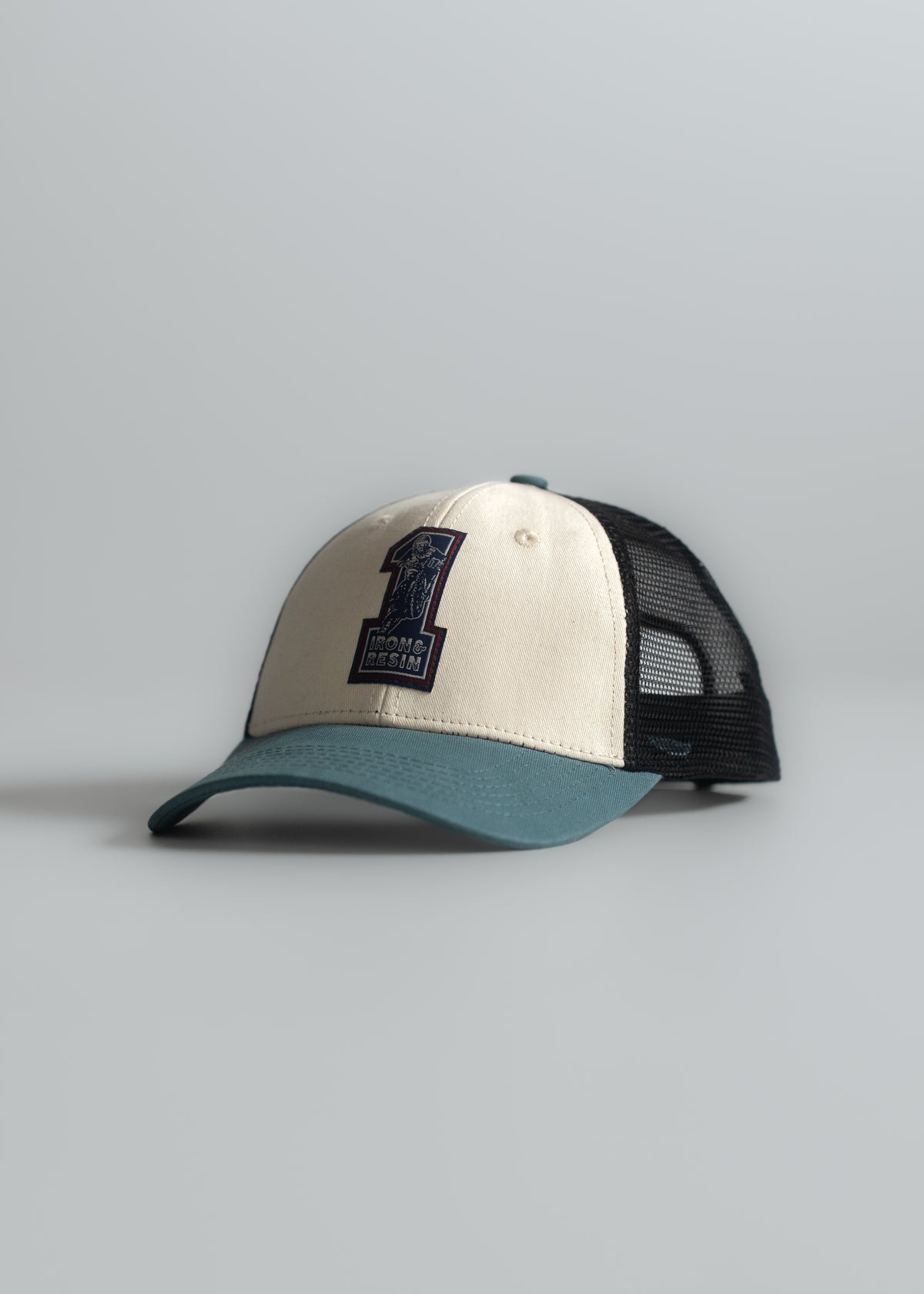 A beige and teal trucker hat with a black mesh back, featuring a large "1" logo on the front, rests on a light gray surface.
