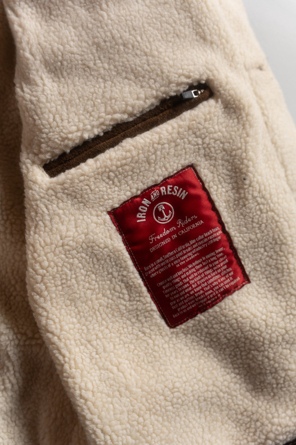 Close-up of a beige fleece jacket with a red "Iron and Resin" label and a brown zippered pocket, showcasing texture and detail.