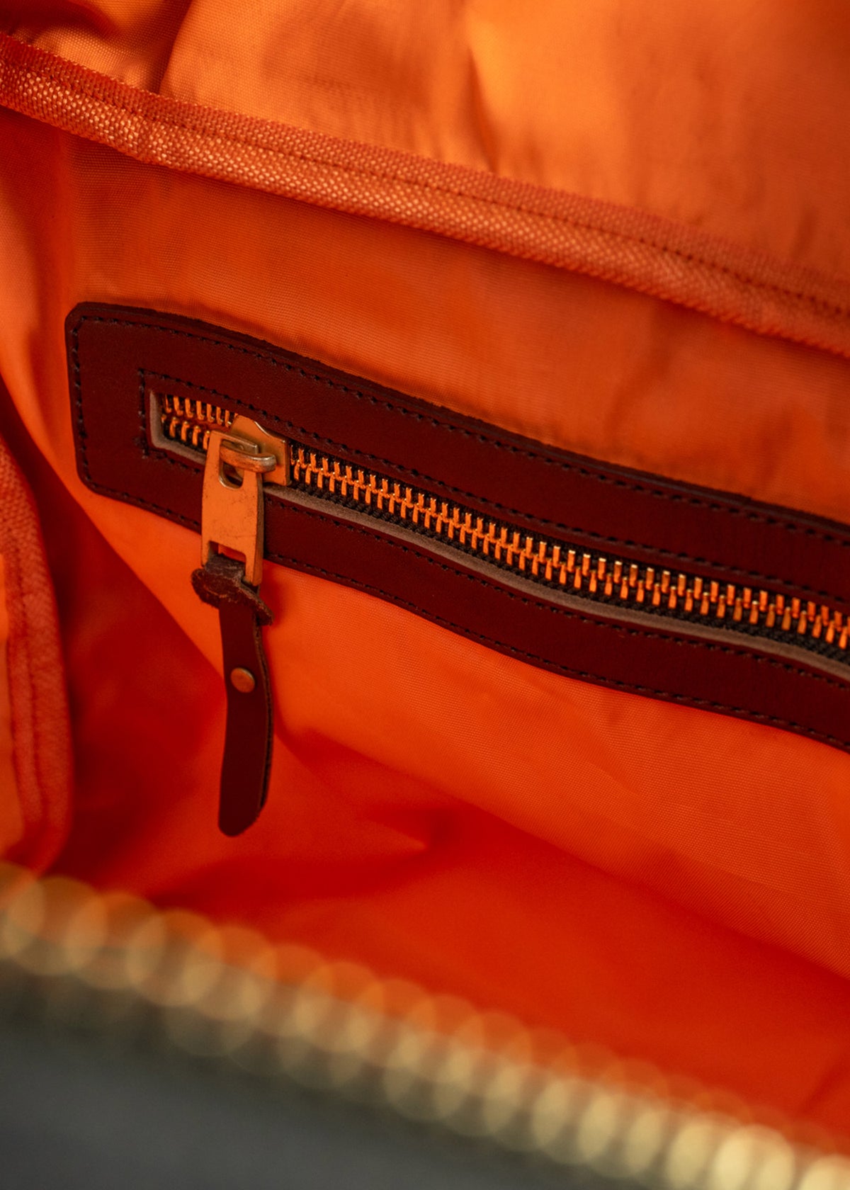Orange fabric bag interior with a brown leather-trimmed zipper pocket, showcasing detailed stitching and texture.