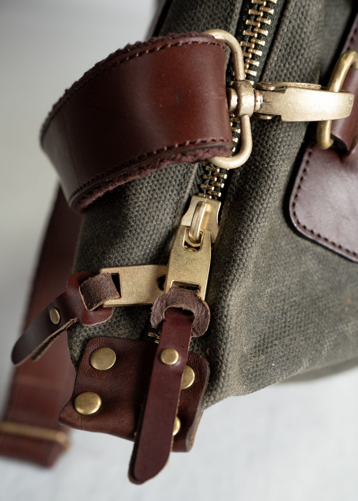 Close-up of a canvas bag's brass zipper and brown leather straps with rivets, highlighting its rugged, vintage design.