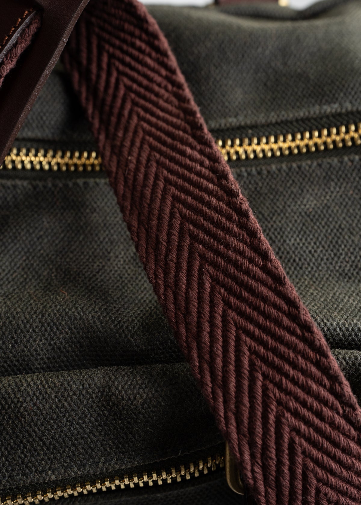 Close-up of a brown woven strap on a dark canvas bag with gold zippers, highlighting texture and detail in the fabric.