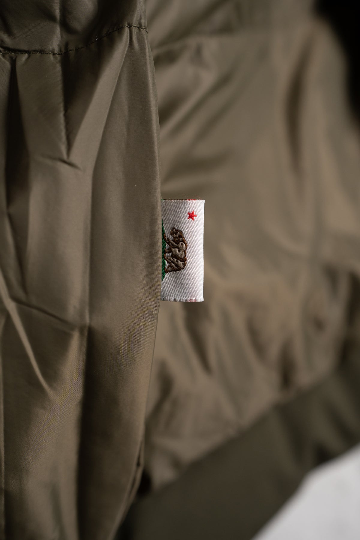 A close-up of a brown jacket's seam with a small white tag featuring a bear and red star, symbolizing California's state flag.