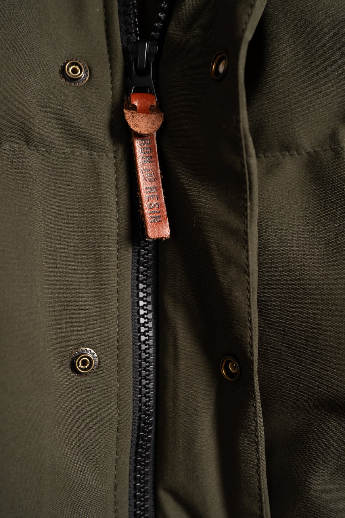 Close-up of an olive jacket's zipper with a leather pull tab. Metal snaps are visible on the fabric, adding detail to the design.