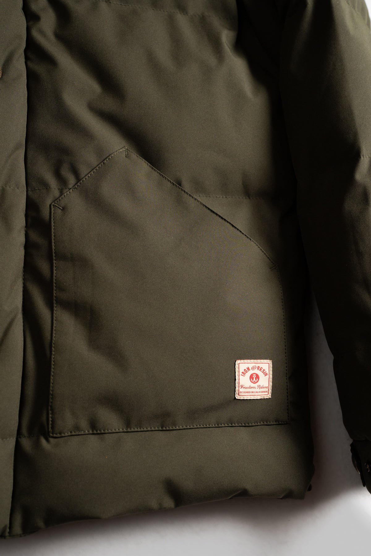 Olive green jacket with a large front pocket and a small red and white label on the bottom right corner, set against a plain backdrop.