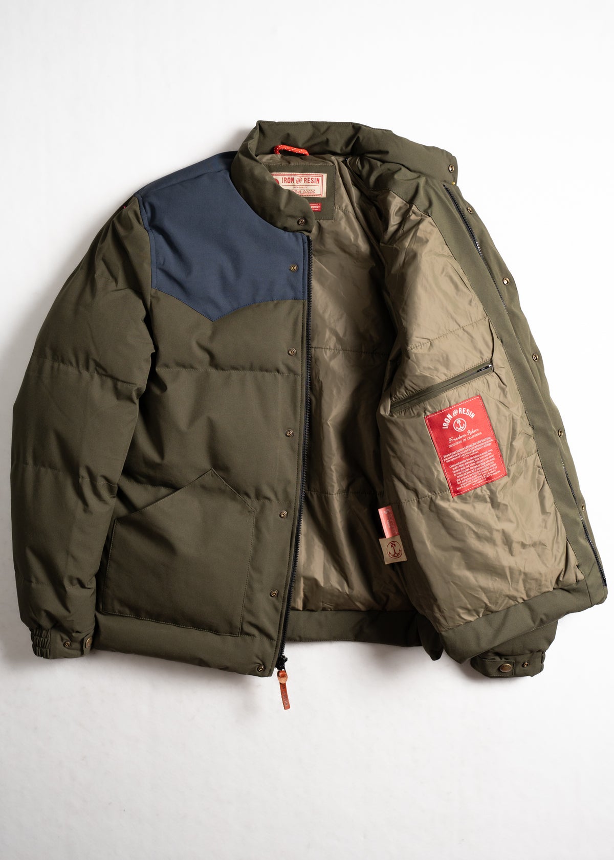 Green puffer jacket with blue shoulder patch, open to reveal beige lining and red label inside. Iron and Resin branding visible.