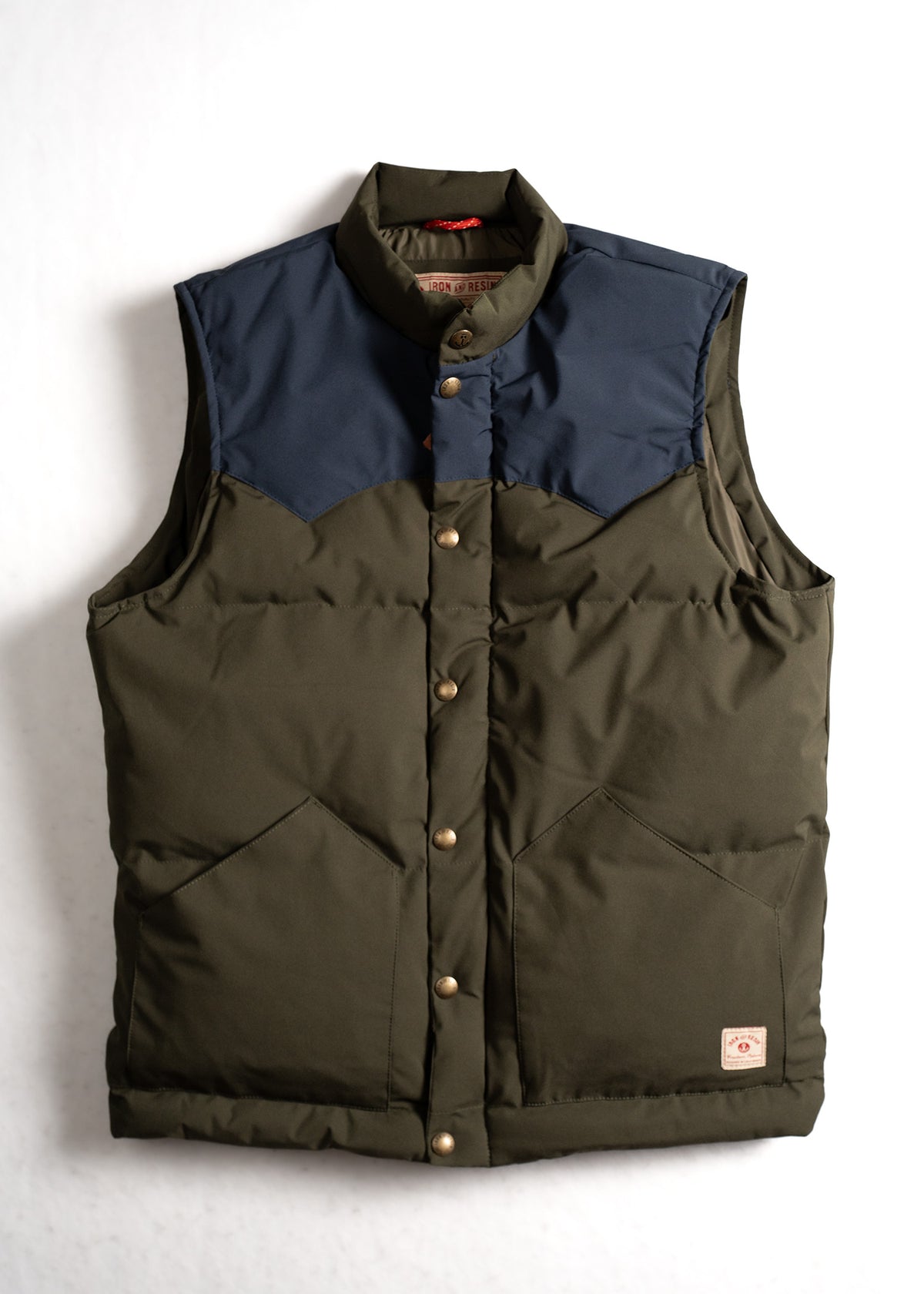 Olive green and navy puffer vest with brass buttons, featuring two front pockets and a high collar, on a white background.