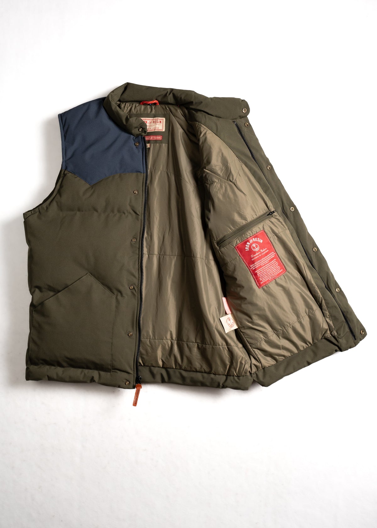 Olive green puffer vest with a navy shoulder, open to reveal a red label inside. Snap buttons and zipper are visible.