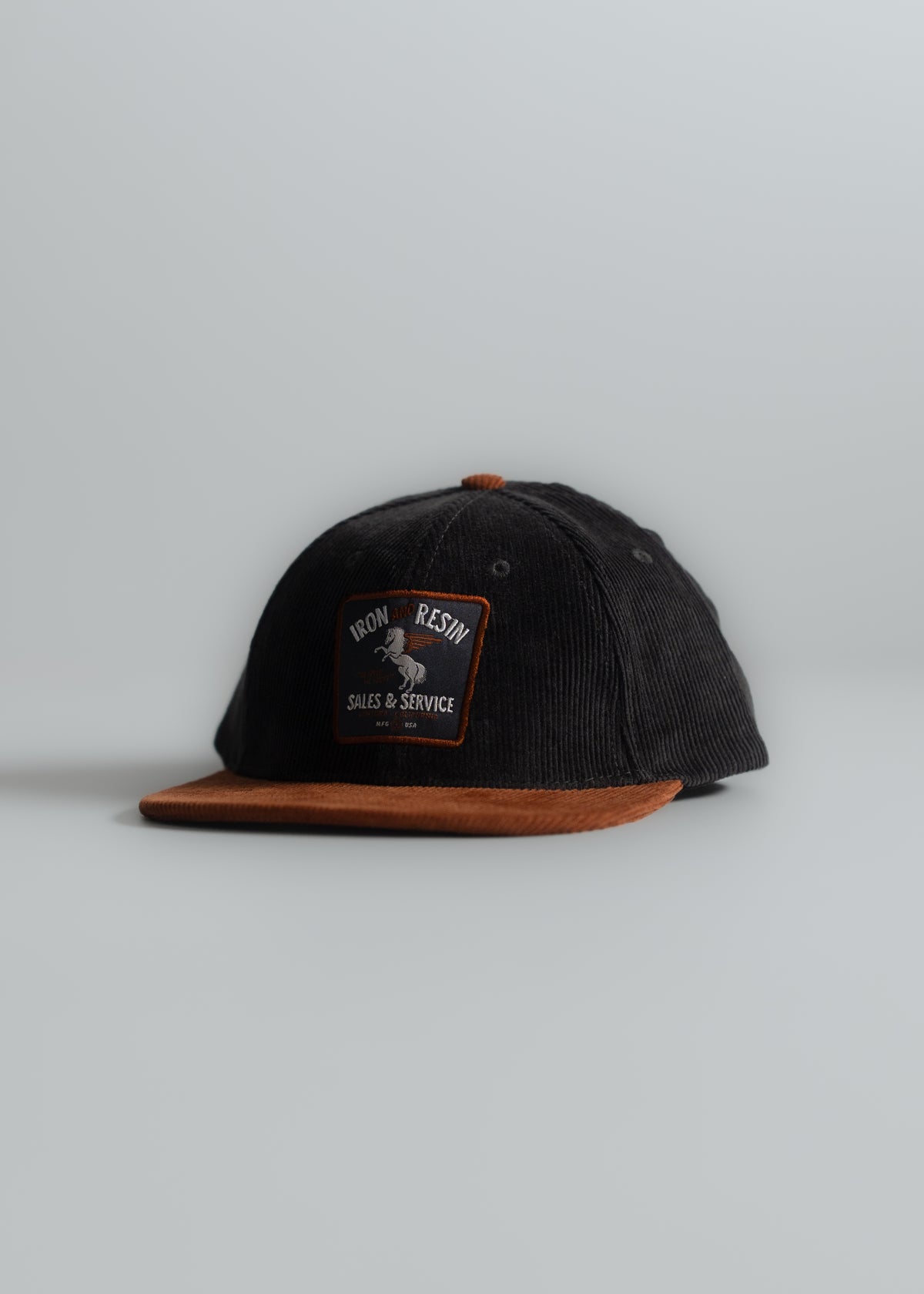 Black cap with brown brim, featuring "Iron and Resin" patch. Simple, stylish design on a light gray background.