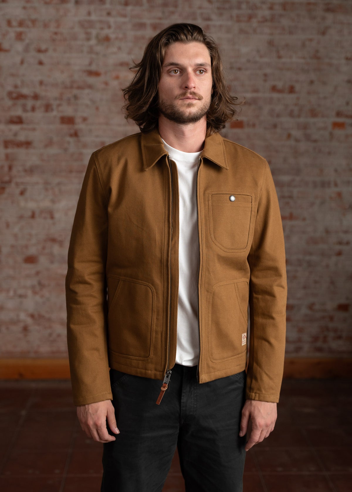 Man in brown jacket and white shirt stands against a brick wall, hands relaxed, with long hair and a thoughtful expression.