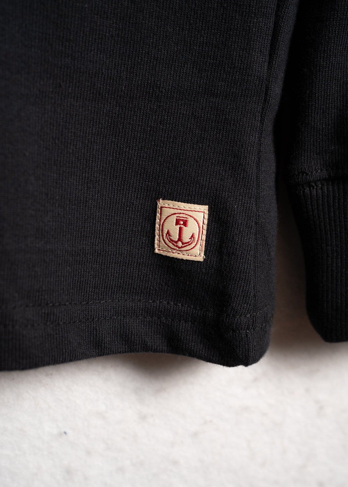 Close-up of a black sweater's hem with a small beige patch featuring a red anchor design, set against a light background.