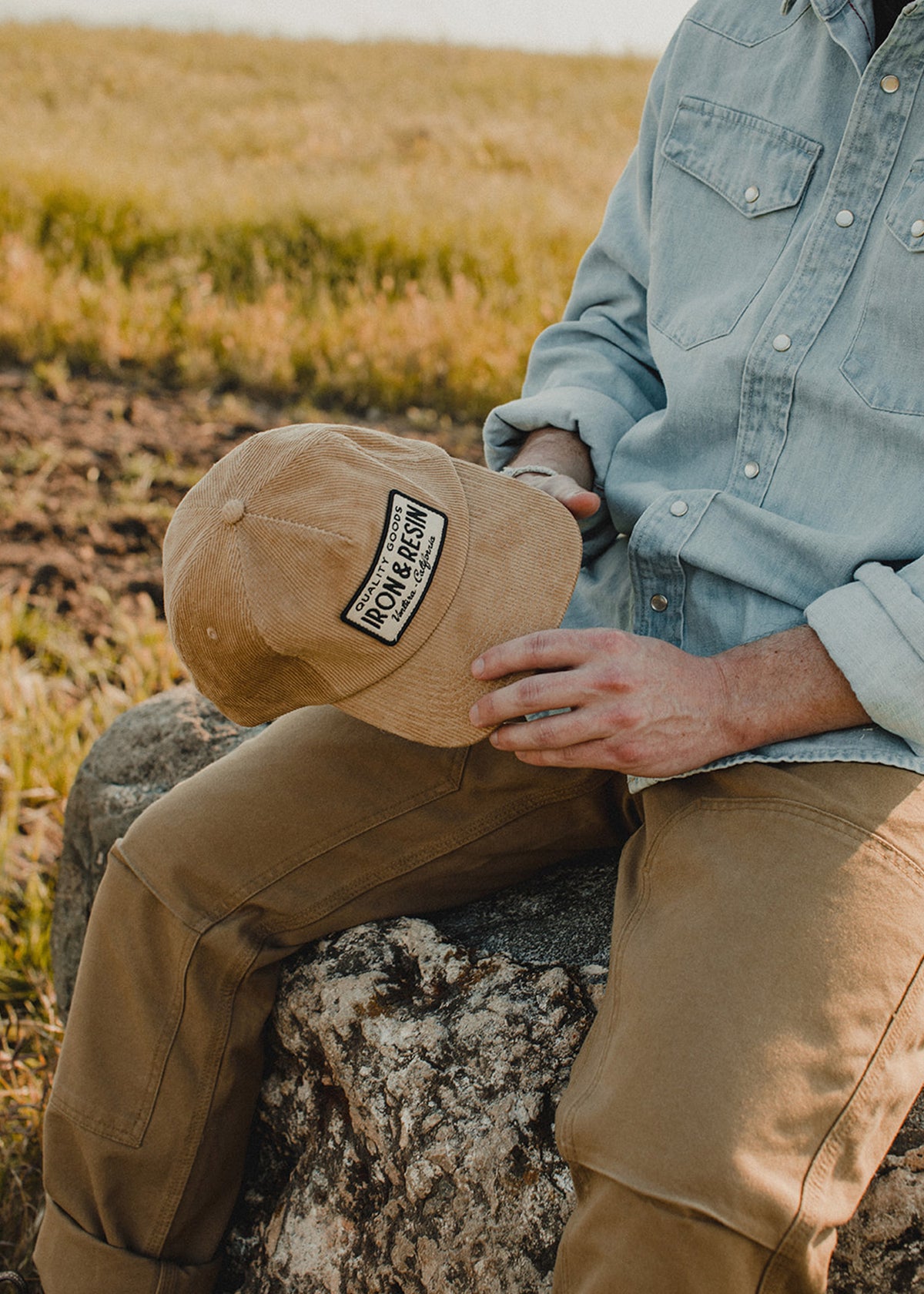Man in denim shirt and brown pants sits on a rock, holding a tan cap labeled "Iron & Resin" in a grassy field.