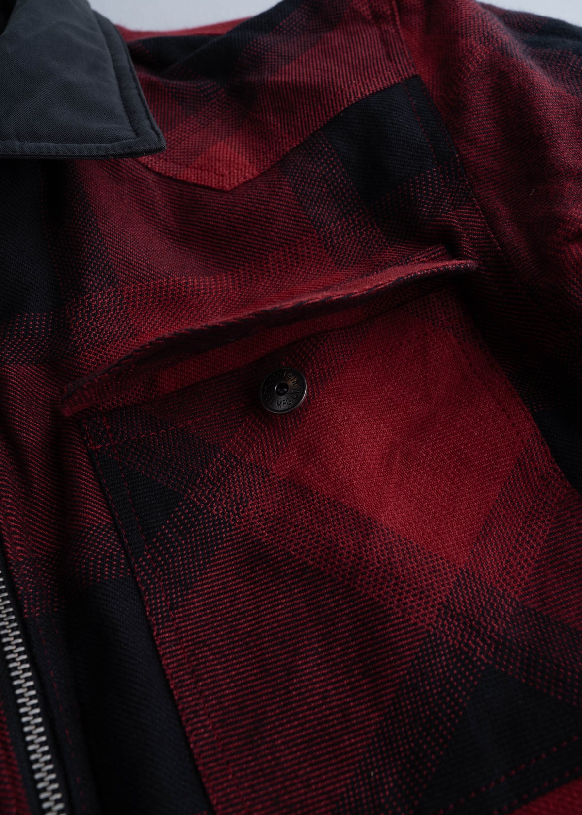 Close-up of a red and black plaid jacket with a buttoned pocket and visible zipper, showcasing its textured fabric.