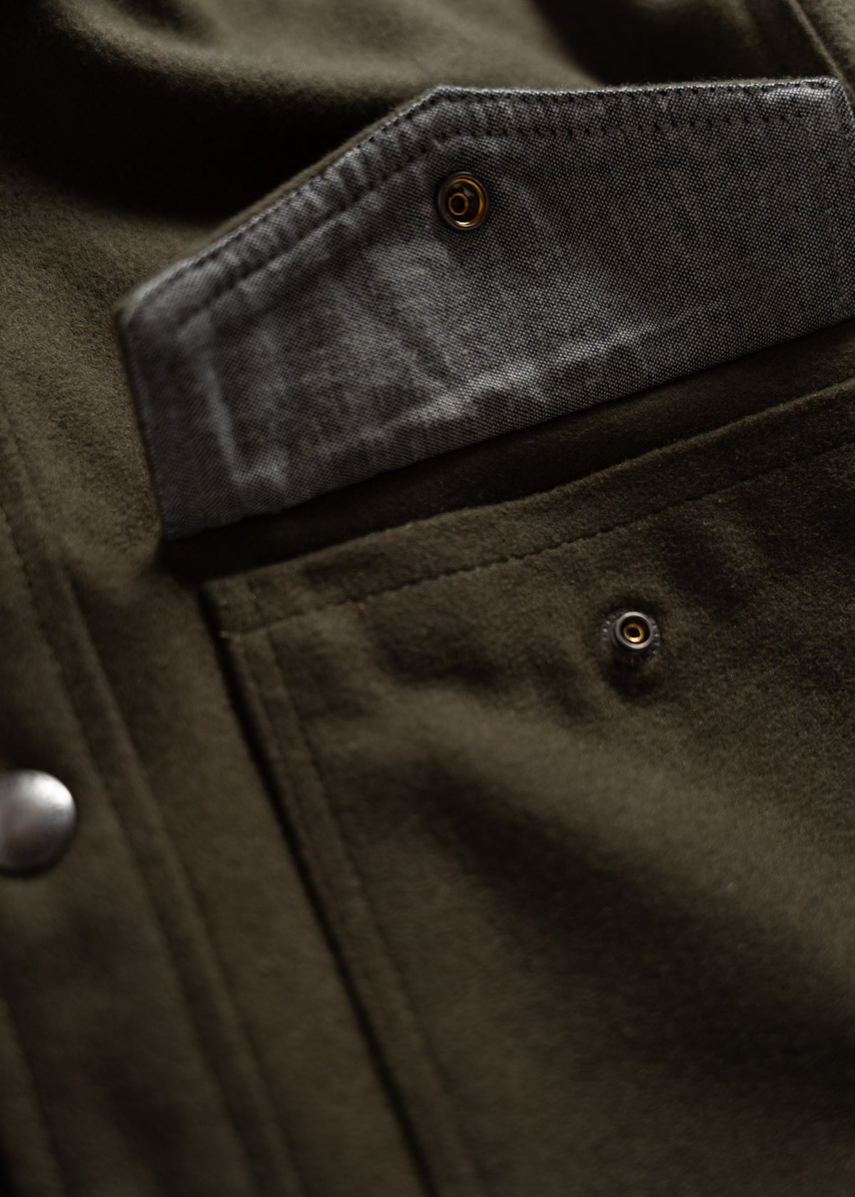 Close-up of a dark green jacket pocket with a gray flap and button detail, highlighting texture and craftsmanship.