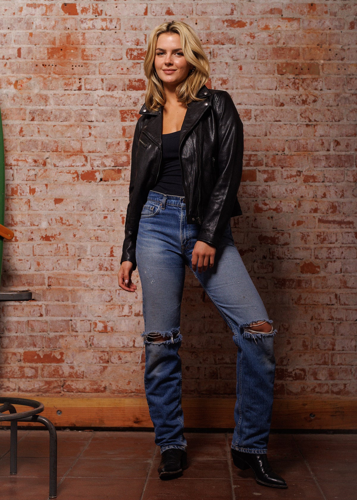 Woman in leather jacket and ripped jeans stands confidently against a rustic brick wall, exuding a casual, stylish vibe.
