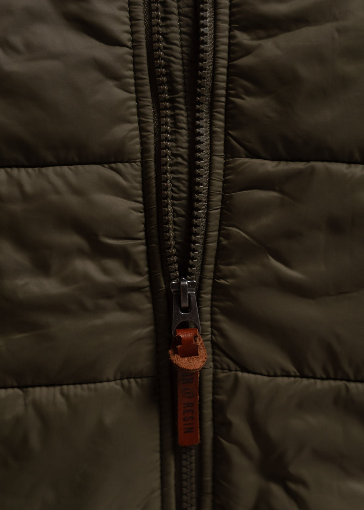 Close-up of a brown quilted jacket with a central zipper and leather pull tab, emphasizing texture and stitching details.