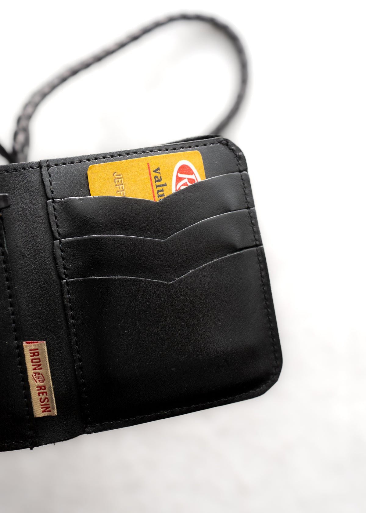 Black wallet with a braided strap, holding a yellow card labeled "Value." Tag reads "Iron and Resin." White background.