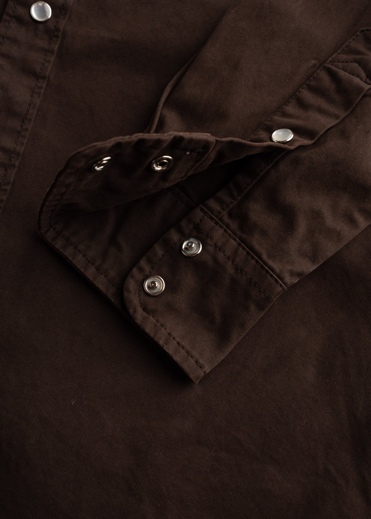Close-up of a brown shirt sleeve with silver snap buttons, showcasing detailed stitching and fabric texture.