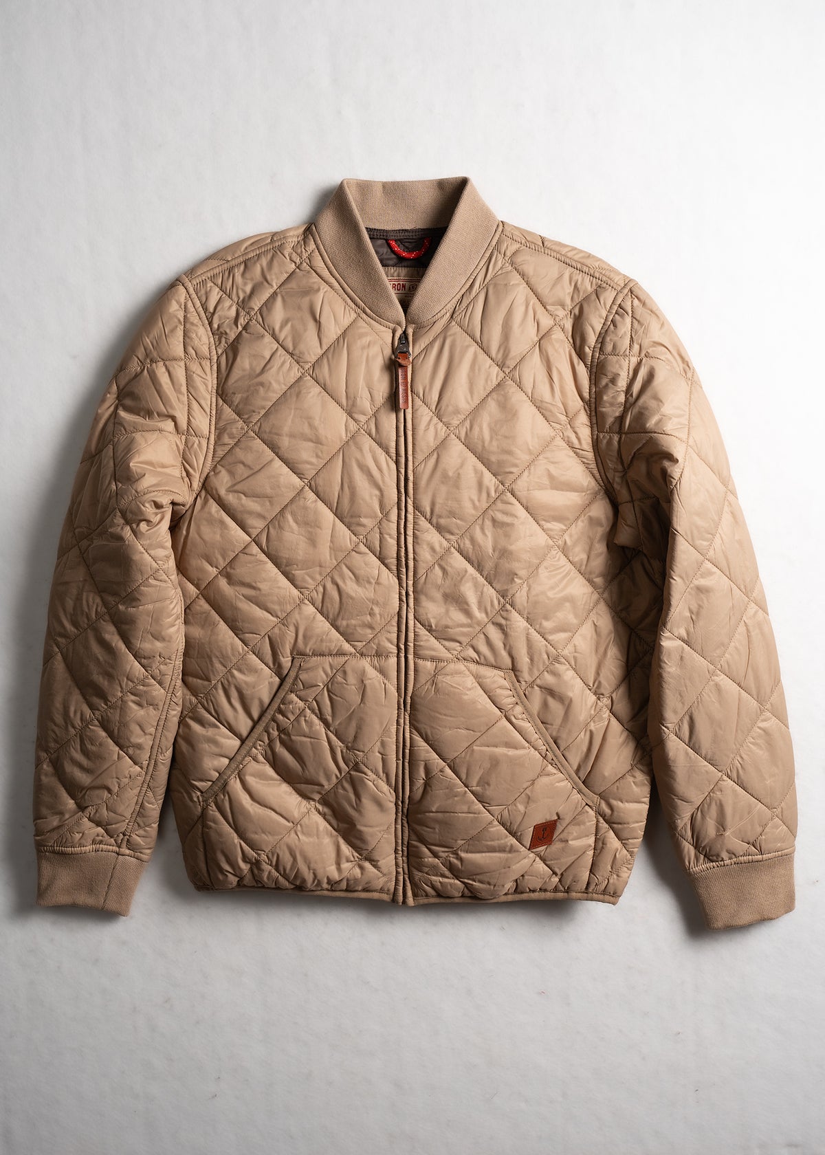Tan quilted bomber jacket with ribbed collar and cuffs, featuring a front zipper and diamond pattern, laid flat on a white surface.