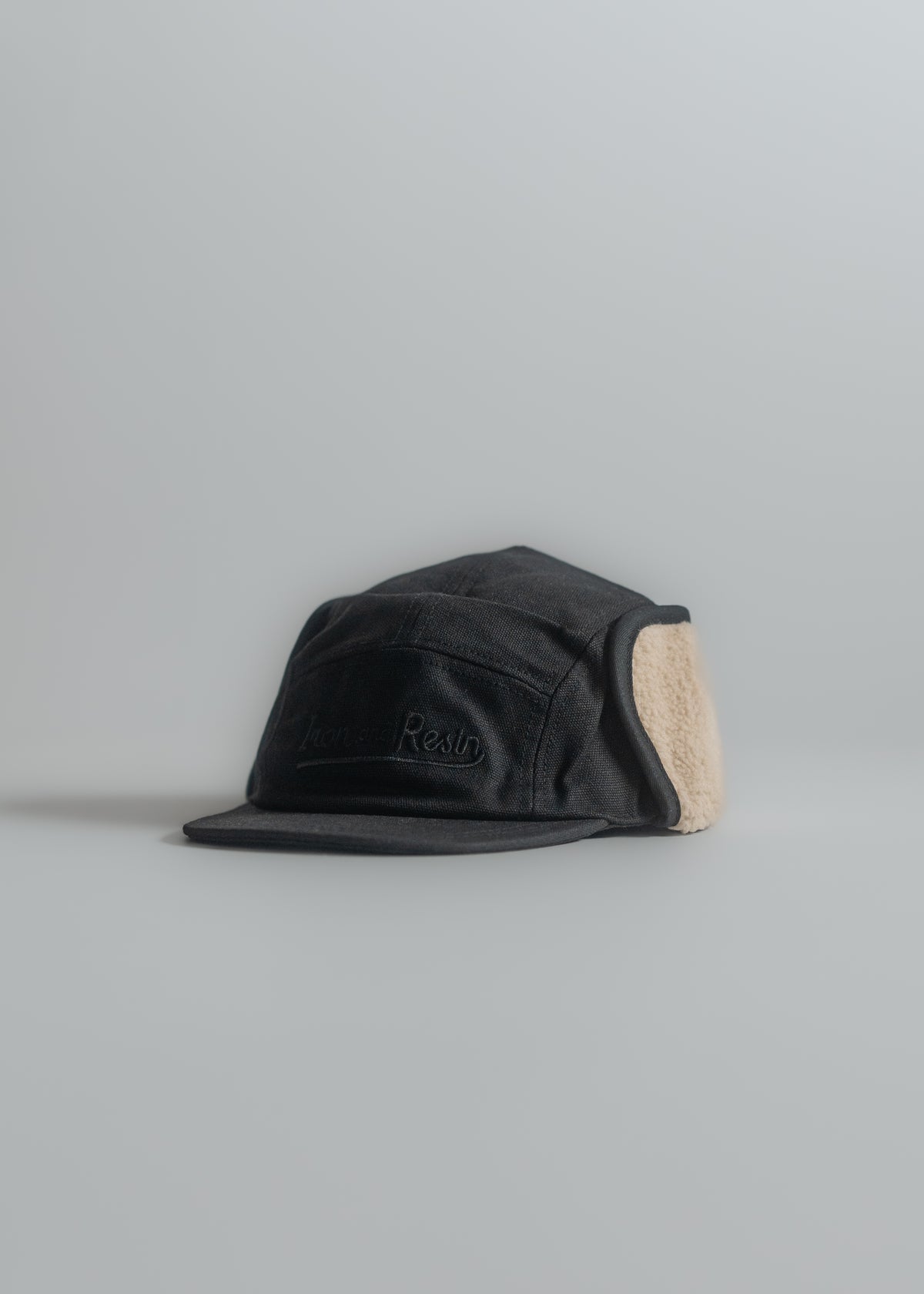 Black cap with ear flaps and beige lining on a gray background, featuring embroidered text on the front panel.