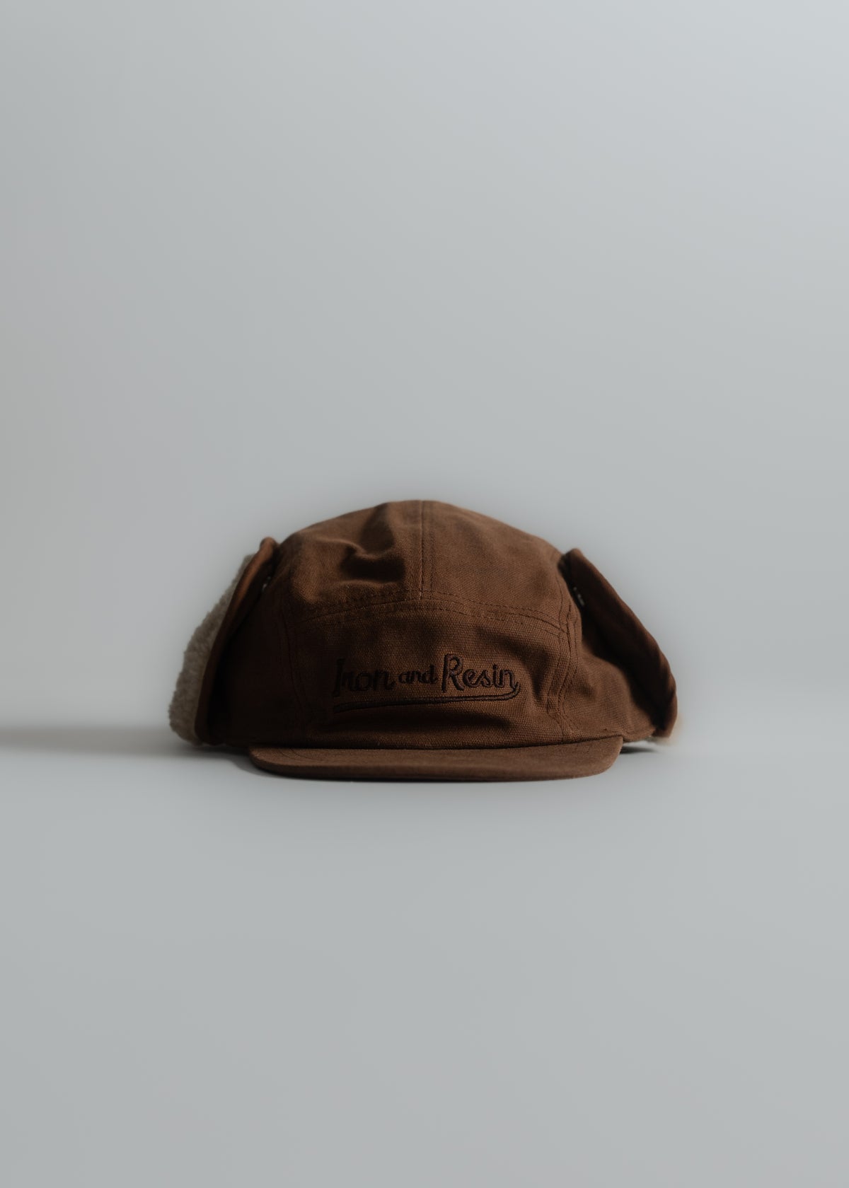 Brown cap with ear flaps, "Iron and Resin" logo, on a light gray background. Simple, rustic design.