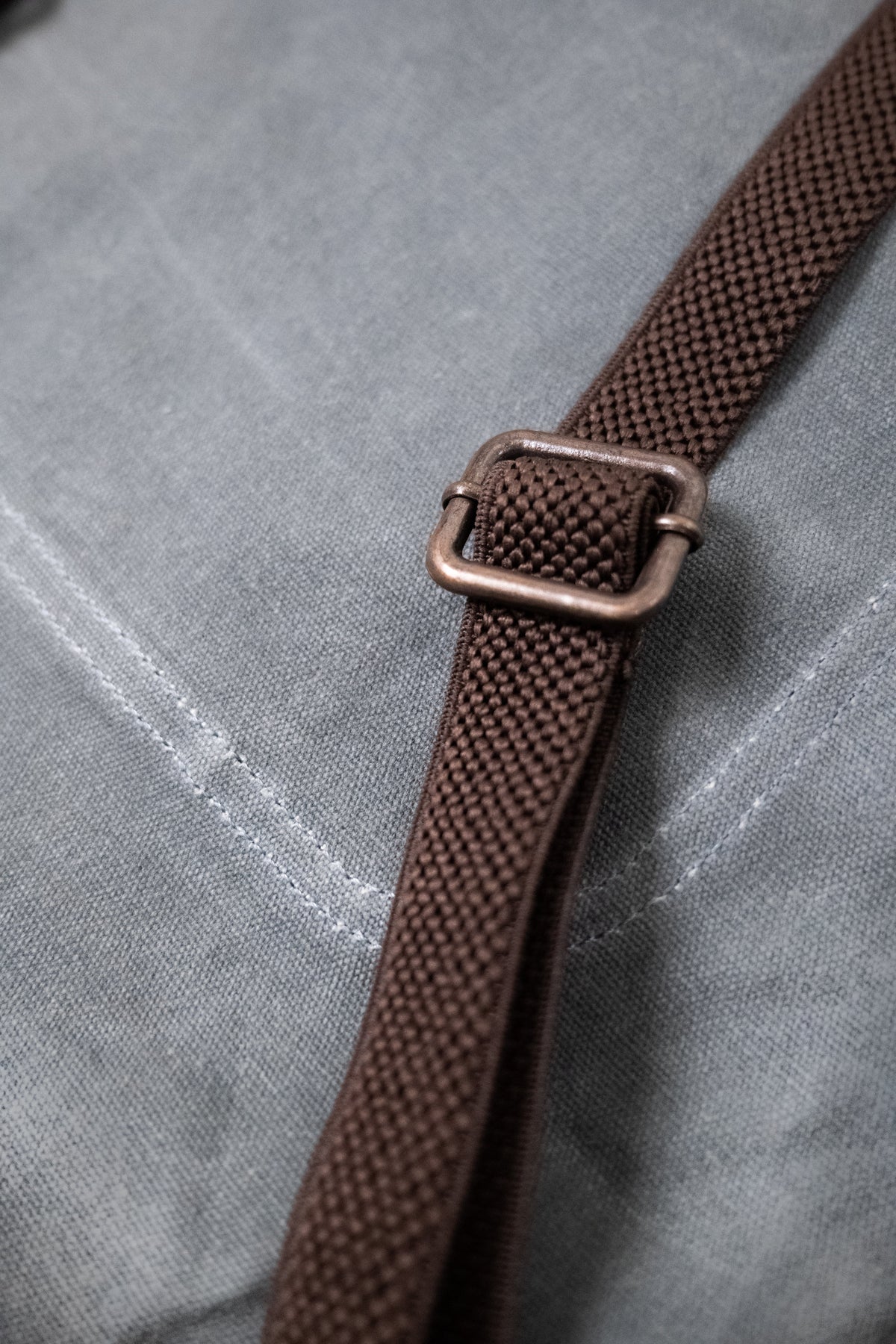 Close-up of a brown strap with a metal buckle on a gray fabric background, highlighting texture and stitching details.
