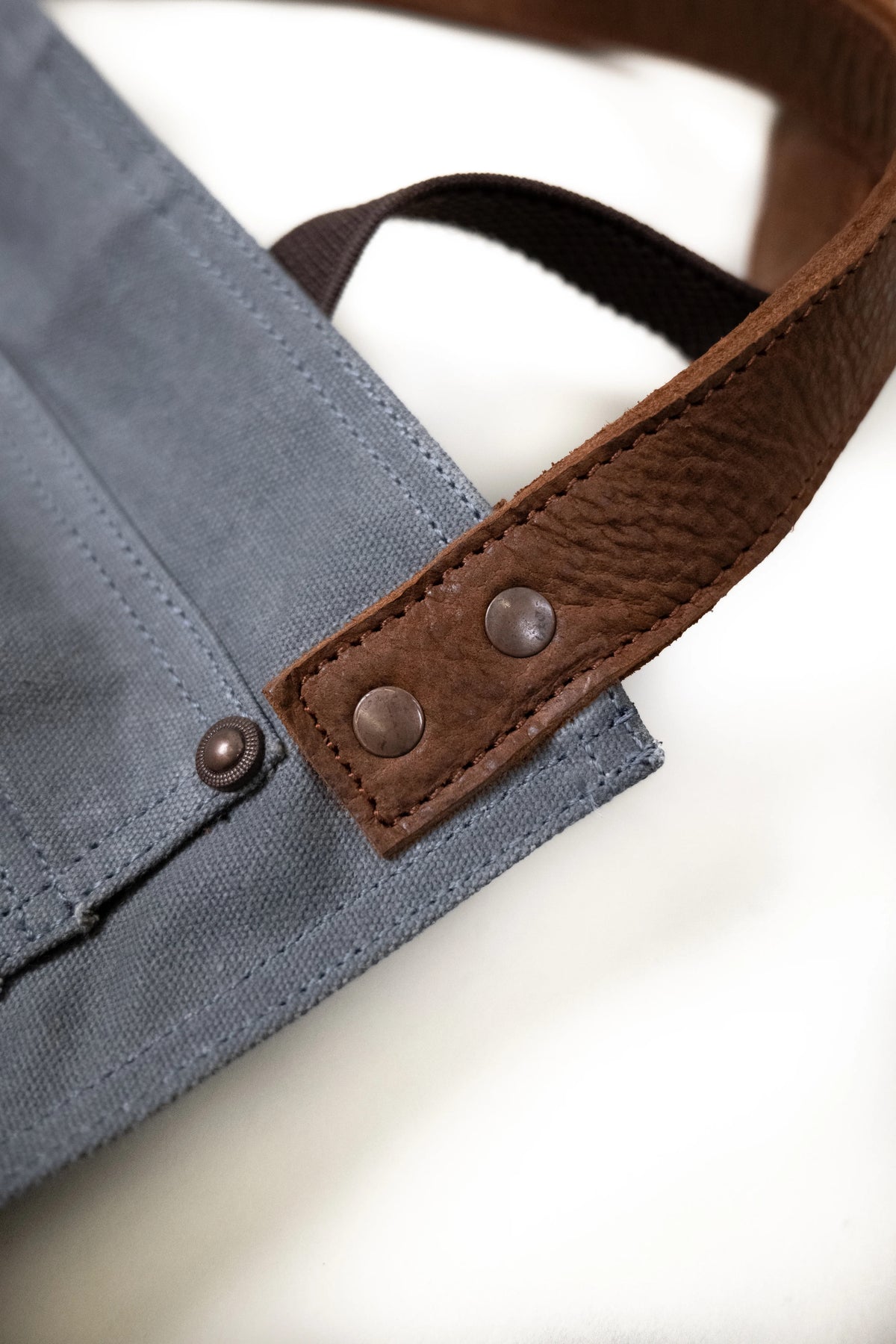 Close-up of a blue canvas bag with brown leather strap and metal rivets, showcasing detailed stitching and texture.