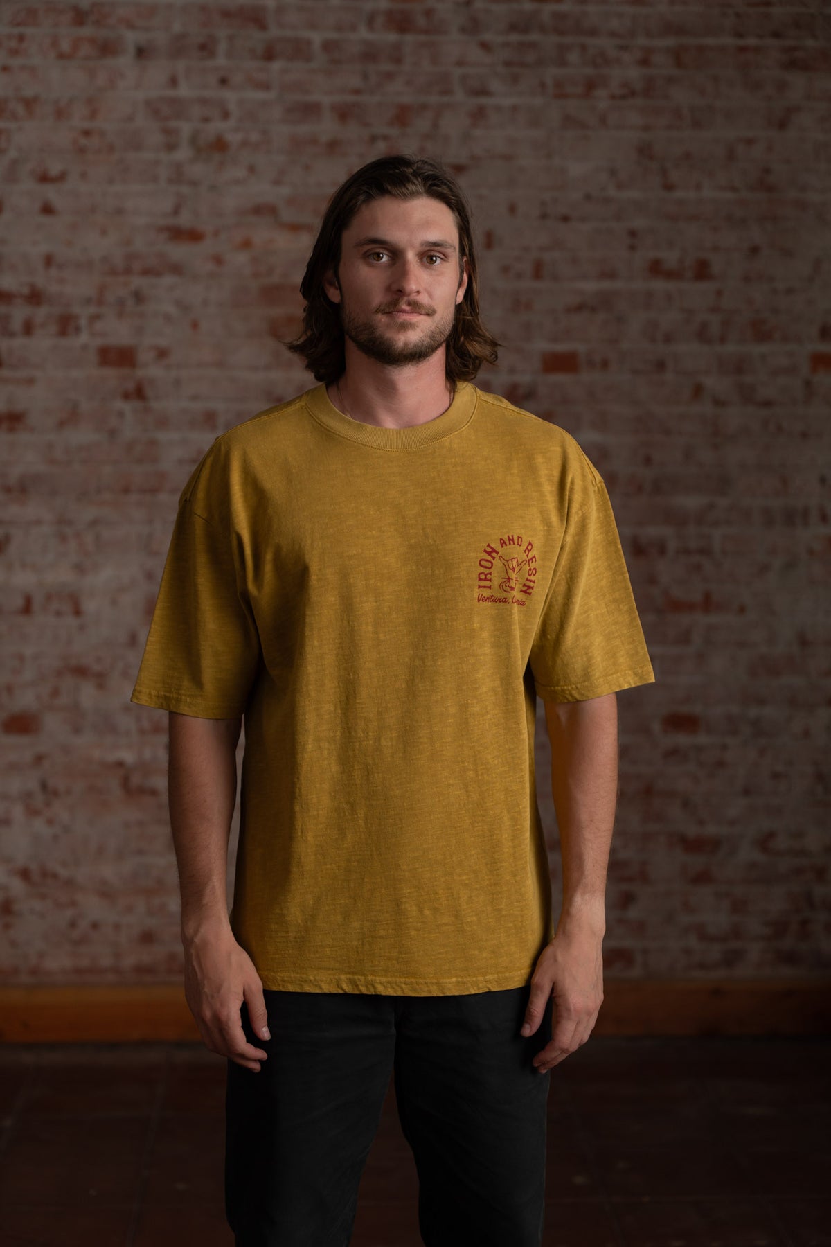Man in mustard t-shirt with "Iron and Resin" logo stands against a textured brick wall, looking directly at the camera.