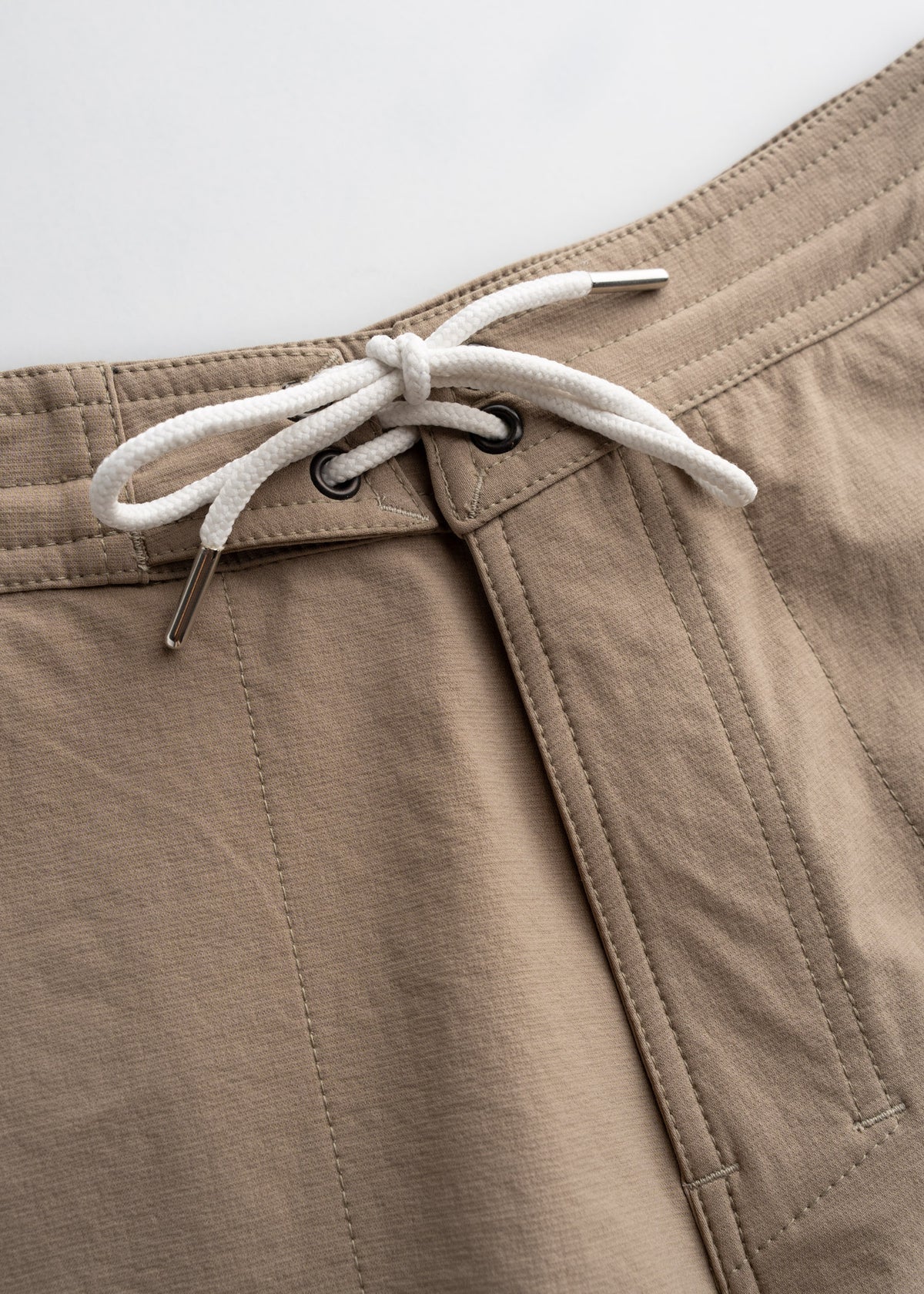 Close-up of beige shorts with a white drawstring and metal eyelets, highlighting the fabric texture and stitching details.